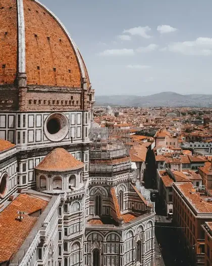 The large, red-tiled dome of a cathedral overlooks a cityscape of red-roofed buildings and distant mountains.