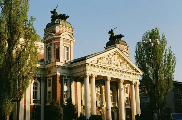 La façade ensoleillée d'un bâtiment néoclassique orné, avec de grandes colonnes blanches, un fronton doré et des statues sur ses tours.