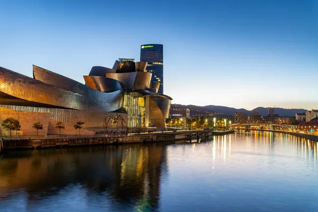 Un moderno museo metallico si erge sulla riva di un fiume al tramonto, le sue luci e lo skyline della città si riflettono nell'acqua.