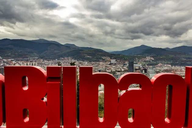 Un grande cartello rosso 'Bilbao' in primo piano con una vista panoramica della città e delle montagne circostanti sotto un cielo nuvoloso.