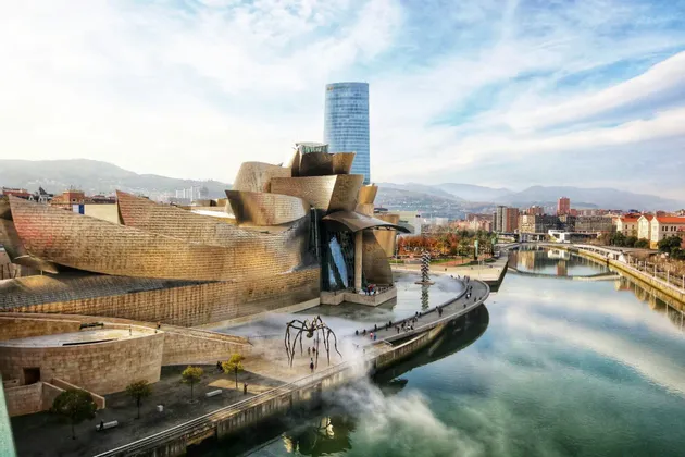 Un edificio metallico astratto con una scultura di ragno gigante sorge lungo un fiume, con la città e le colline visibili sullo sfondo.