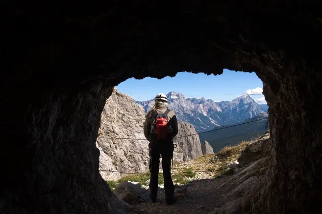 Un escursionista con un casco e uno zaino rosso si trova all'imboccatura di una grotta, guardando un'ampia catena montuosa illuminata dal sole.