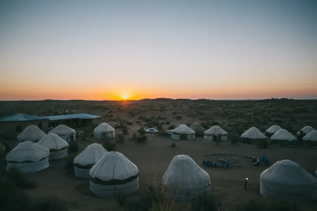 Ein Lager traditioneller Jurten erstreckt sich bei Sonnenuntergang über eine Wüstenlandschaft, die Sonne glüht am Horizont.