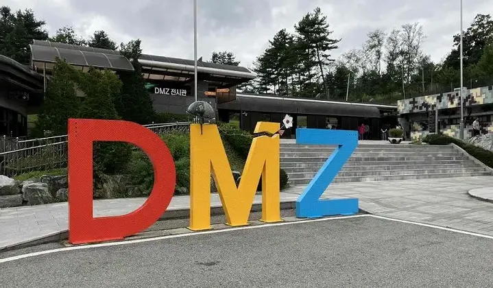 Grandi lettere colorate che compongono la scritta DMZ si ergono di fronte a un padiglione espositivo, con un fiore bianco che spunta dalla 'M' spezzata.