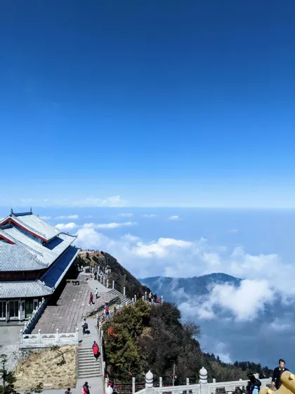 Veduta dall'alto di un edificio tradizionale in cima a una montagna, con persone che percorrono un sentiero affacciato su un mare di nuvole e un vasto cielo blu.