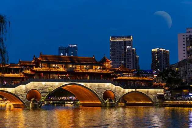 Un ponte tradizionale cinese illuminato con edifici in cima attraversa un fiume di notte, con moderni grattacieli sullo sfondo sotto una luna crescente.