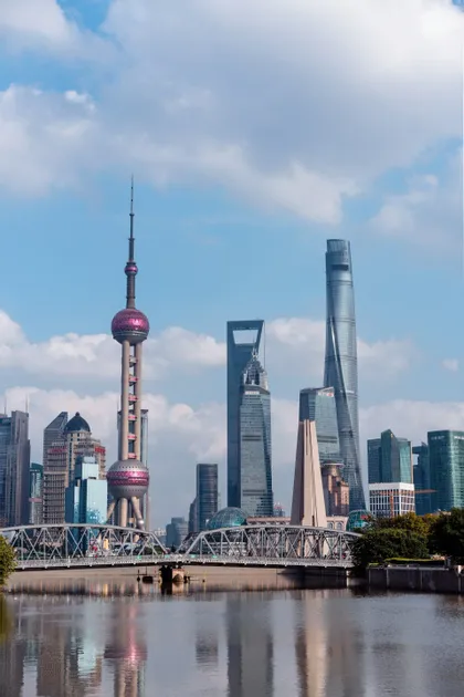 Lo skyline moderno di Shanghai, con la Torre della Perla Orientale, si erge sopra un fiume e un ponte d'acciaio sotto un cielo blu con nuvole bianche.