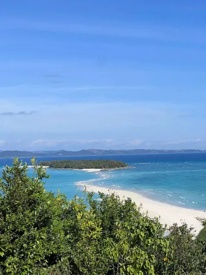 Una vista dall'alto su un fogliame verde che mostra una lingua di sabbia bianca che conduce a un'isola tropicale lussureggiante circondata da acqua turchese.