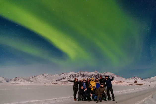 Un grupo de WeRoad posa para una foto bajo la aurora boreal verde en un paisaje nevado con montañas.