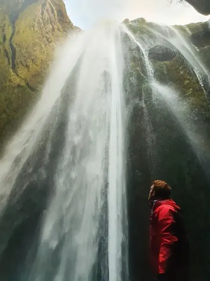 Eine Person in roter Jacke steht am Fuße eines imposanten Wasserfalls und blickt auf das Wasser, das eine moosbewachsene Klippe herabstürzt.