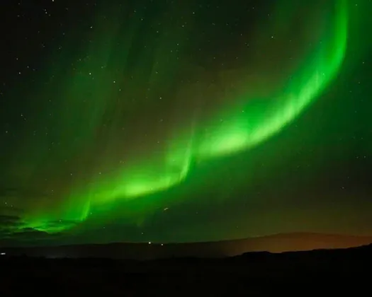 Una aurora boreal verde brillante se arquea en un cielo nocturno estrellado sobre la silueta de un paisaje oscuro.