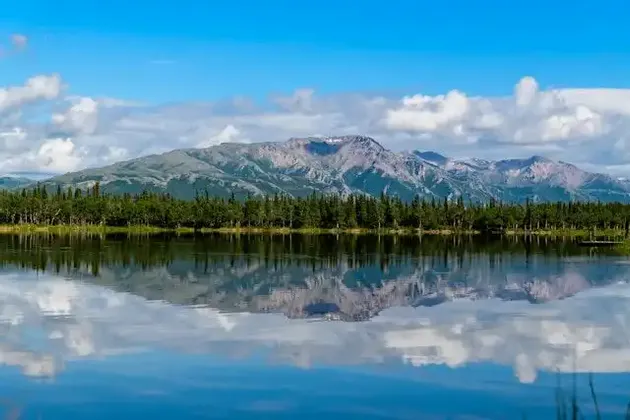 A mountain range and a line of trees reflecting on the calm surface of a lake under a partly cloudy sky.