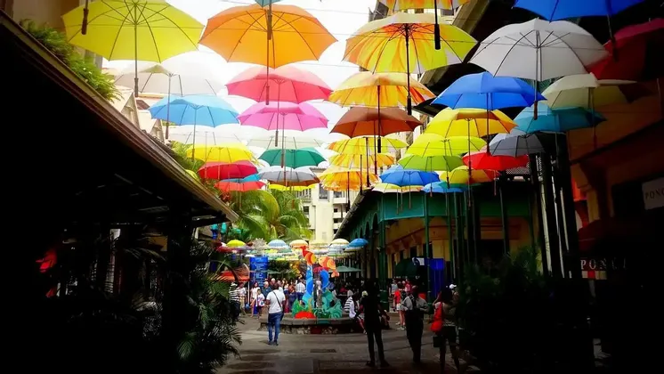 Un baldacchino di ombrelli multicolori pende su una vivace strada cittadina piena di pedoni e negozi.