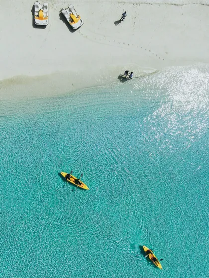 Vista aerea di due kayak gialli in acque limpide e turchesi accanto a una spiaggia di sabbia bianca con persone e pedalò.