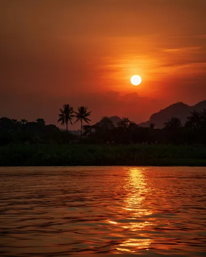 Il sole tramonta in un cielo arancione intenso, la sua luce si riflette su un fiume con sagome di palme e colline sulla riva opposta.