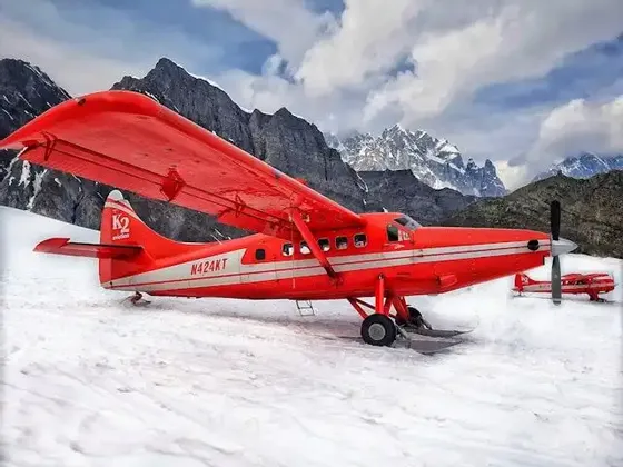 A red propeller plane fitted with skis is parked on a snowy glacier, with a rugged mountain range in the background.