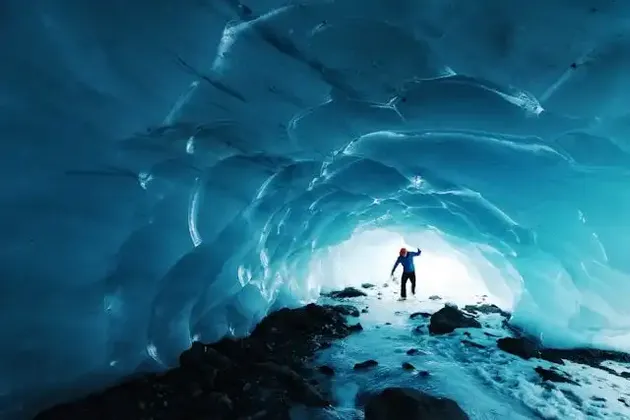 A person in a blue jacket and red hat stands inside a glowing blue ice cave, looking towards the bright entrance.