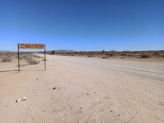 Un cartello 'Tropico del Capricorno' si erge accanto a una lunga strada sterrata in un vasto paesaggio desertico sotto un cielo azzurro.