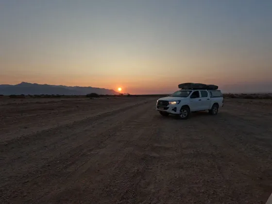 Un pick-up bianco con tenda da tetto è parcheggiato su una strada sterrata in un vasto paesaggio desertico mentre il sole tramonta dietro le montagne.