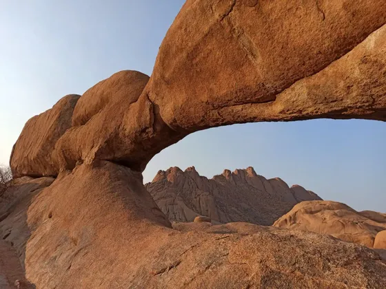 Un imponente arco naturale di formazioni rocciose rossastre, con vista su montagne lontane attraverso l'apertura sotto un cielo sereno.