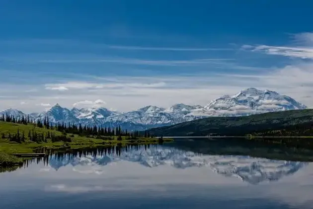 A snow-capped mountain range and a green treeline are perfectly reflected in the calm waters of a lake under a blue sky.