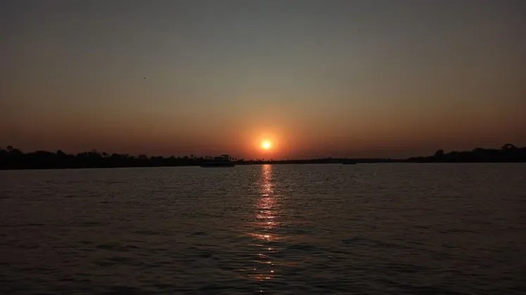 Il sole tramonta su un ampio specchio d'acqua, la sua luce arancione si riflette sulla superficie, con una barca visibile in lontananza.