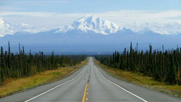 A straight paved road stretches through a dense evergreen forest towards a large, snow-covered mountain range under a partly cloudy sky.
