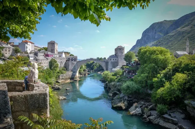 Una persona con i capelli biondi siede su un muro di pietra che si affaccia su una città storica con un ponte ad arco in pietra che attraversa un fiume turchese.