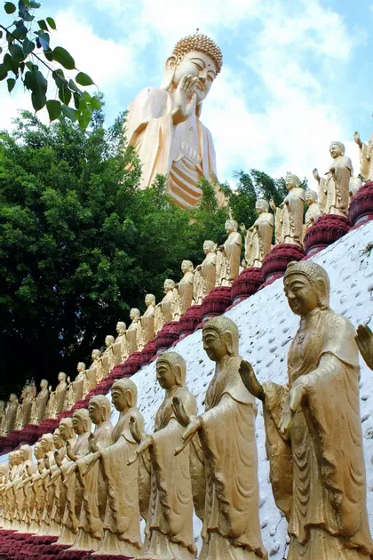 Eine riesige goldene Buddha-Statue thront über vielen kleineren goldenen Statuen, die in Reihen an einem Berghang unter einem teilweise bewölkten Himmel angeordnet sind.