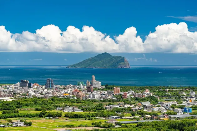 Eine Küstenstadt, umgeben von Grün, blickt auf eine große Insel im Meer unter blauem Himmel mit bauschigen weißen Wolken.