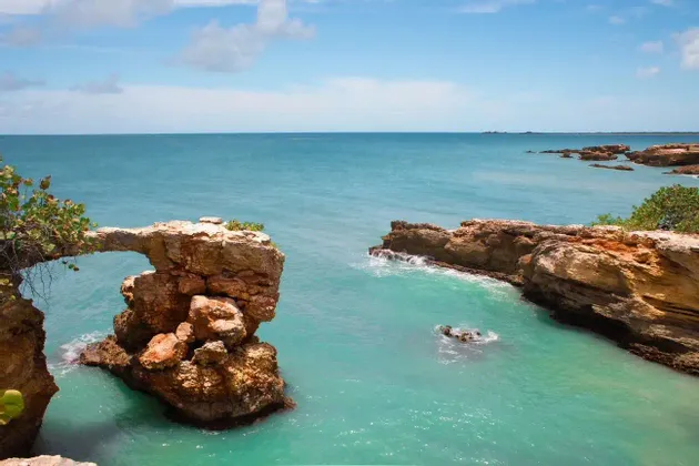 A natural rock arch formation extends over turquoise coastal waters with a blue sky and clouds above.