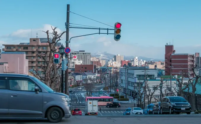 Una vista ampia di una strada cittadina giapponese con traffico, edifici e montagne innevate distanti sotto un cielo azzurro chiaro.