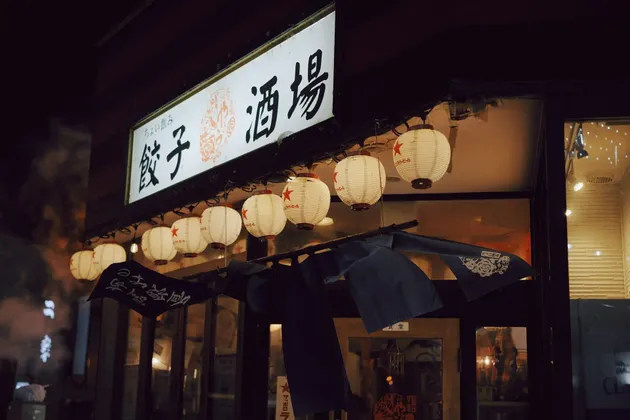 Lanterne di carta bianche e illuminate sono appese in fila sotto un'insegna luminosa con caratteri giapponesi, a indicare un ingresso di notte.
