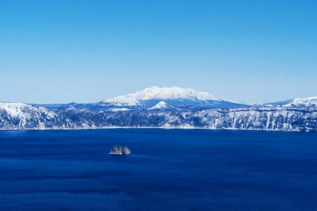 Un'ampia vista di un lago blu profondo con un piccolo isolotto roccioso, sullo sfondo montagne innevate sotto un cielo limpido.