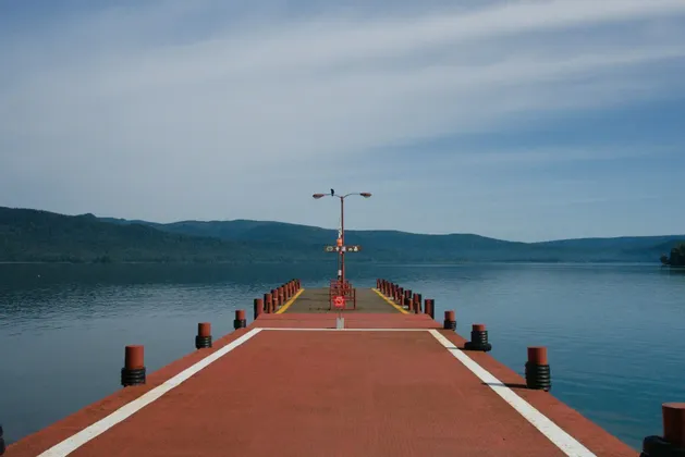 Un lungo molo rosso si estende su un lago tranquillo verso lontane montagne boscose sotto un cielo blu.
