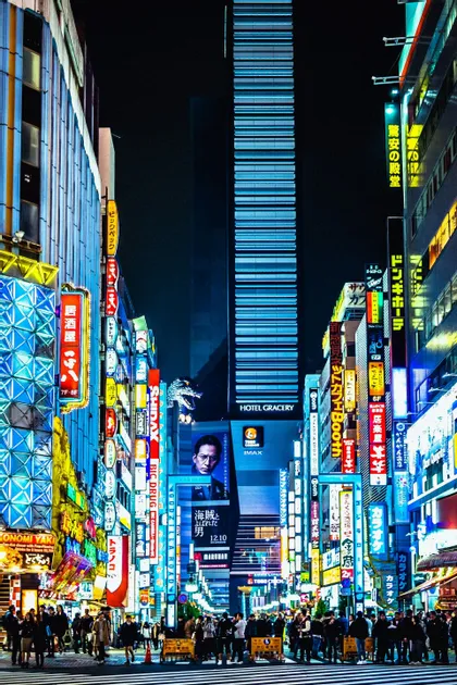 Una strada cittadina animata di notte, affollata di persone e illuminata da vivaci insegne al neon su edifici imponenti.