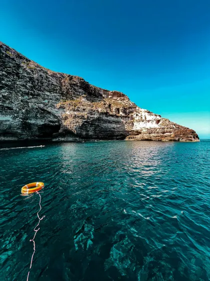 Un salvagente arancione galleggia sul mare turchese intenso accanto a una scogliera rocciosa sotto un cielo azzurro limpido.