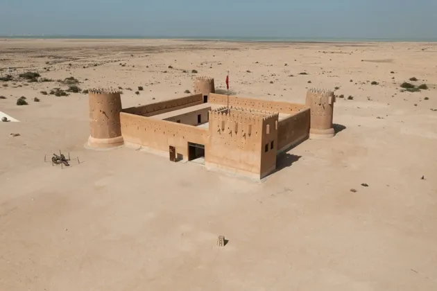 Una ripresa aerea di una fortezza quadrata color sabbia con quattro torri angolari, situata in un vasto deserto con il mare sullo sfondo.