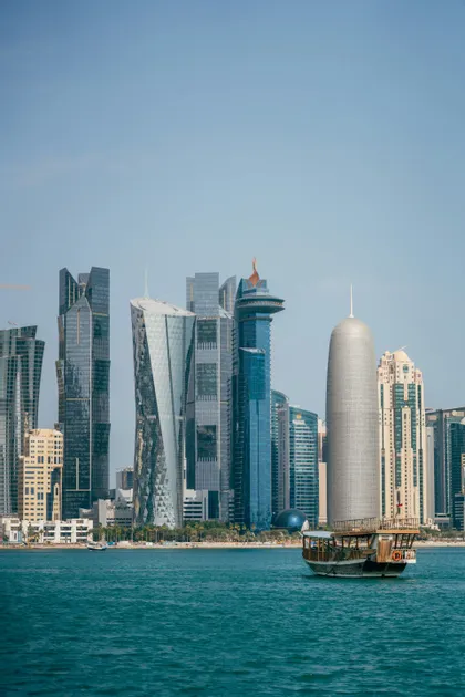 Una barca tradizionale in legno naviga su acque turchesi, con i moderni grattacieli di uno skyline cittadino sullo sfondo.