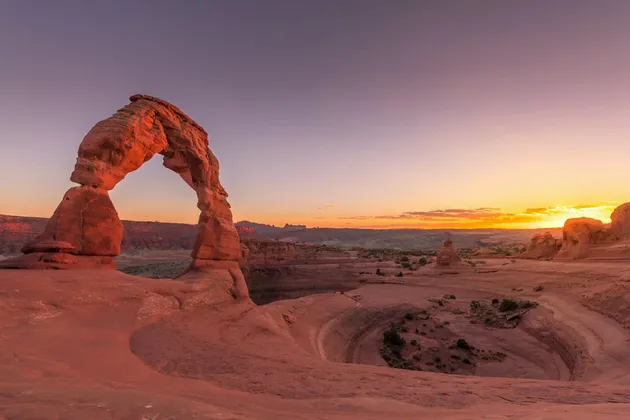 Un grande arco naturale di arenaria risplende alla luce dell'alba su un vasto paesaggio desertico di roccia rossa.