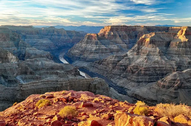 Un vasto canyon stratificato con un fiume sottostante, ammirato da un promontorio roccioso rosso all'alba, sotto un cielo parzialmente nuvoloso.