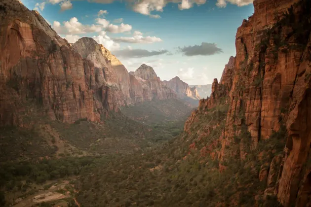 Un'ampia veduta di un canyon di roccia rossa con una rigogliosa valle verde sottostante, sotto un cielo azzurro parzialmente nuvoloso.