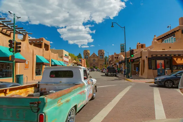 Un pickup vintage turchese su una strada di mattoni fiancheggiata da edifici in stile adobe, con una grande cattedrale visibile alla fine della strada sotto un cielo blu.