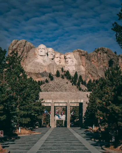 Il Monte Rushmore National Memorial visto dal vialetto d'ingresso in pietra, fiancheggiato da pini sotto un cielo azzurro.