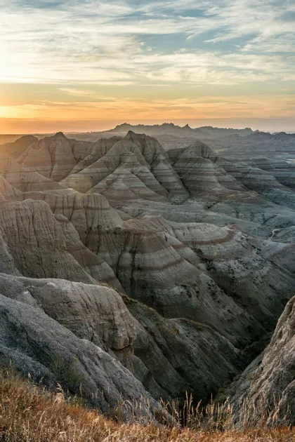Un vasto paesaggio di formazioni rocciose stratificate (calanchi) illuminato dalla calda luce di un tramonto sotto un cielo parzialmente nuvoloso.