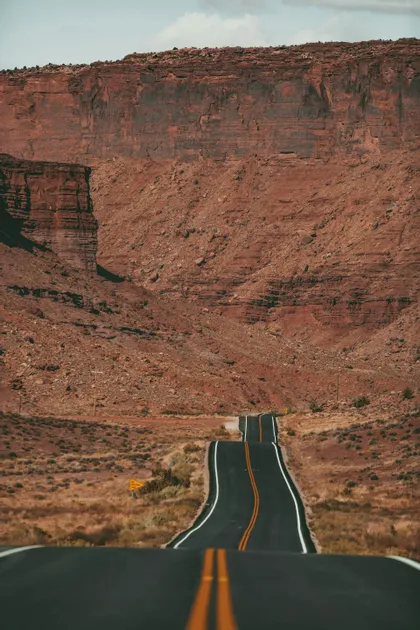 Una strada asfaltata vuota con linee gialle si snoda su e giù attraverso un paesaggio arido verso un'imponente parete di canyon roccioso rosso.