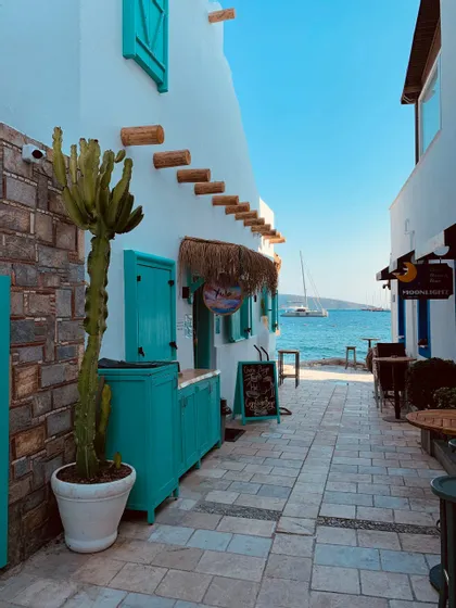 Uno stretto vicolo in pietra tra edifici imbiancati con dettagli turchesi conduce a un mare blu con barche all'orizzonte.