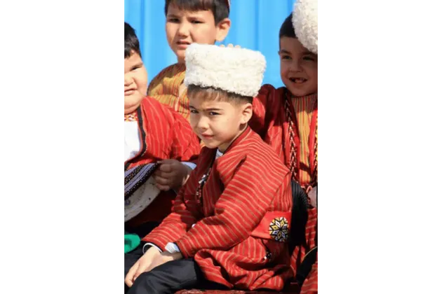 Un bambino, con una giacca tradizionale a righe rosse e un cappello di lana bianco, siede in un gruppo di bambini, guardando la fotocamera.