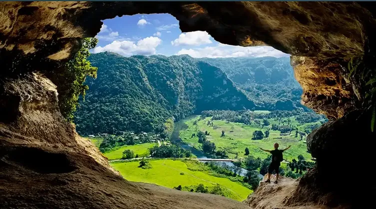 A person with arms outstretched stands at the mouth of a cave, looking out over a lush green valley with mountains and a river.