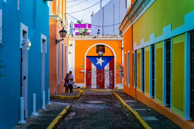 A narrow cobblestone alley flanked by colorful buildings, with a large door at the end painted with the flag of Puerto Rico.